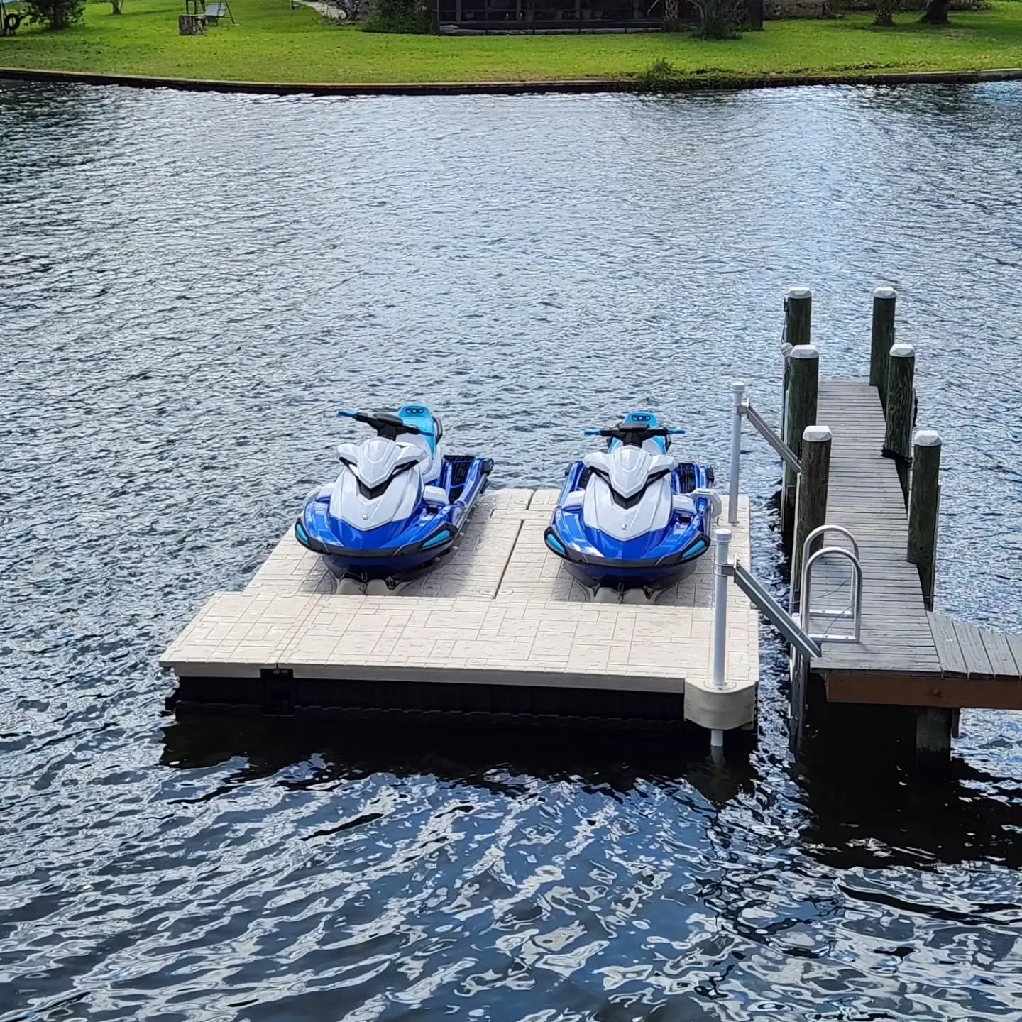 Jet Ski Docks:Two blue and white Yamaha WaveRunners docked side-by-side on a tan modular floating platform. The platform is connected to a narrow wooden walkway extending into a large body of water.