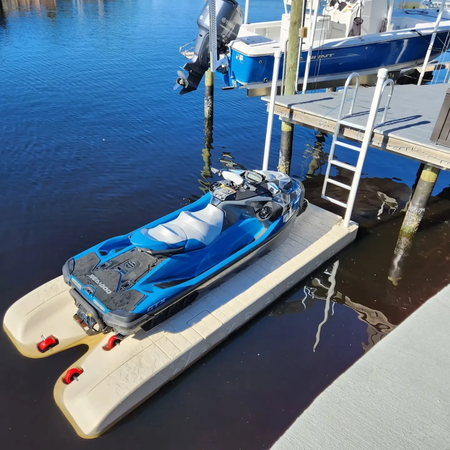 Jet Ski Dock: A blue and grey Sea-Doo jet ski parked on a tan floating PWC port. The port is attached to a wooden dock with a swim ladder in a canal, with a larger boat on a lift in the background.