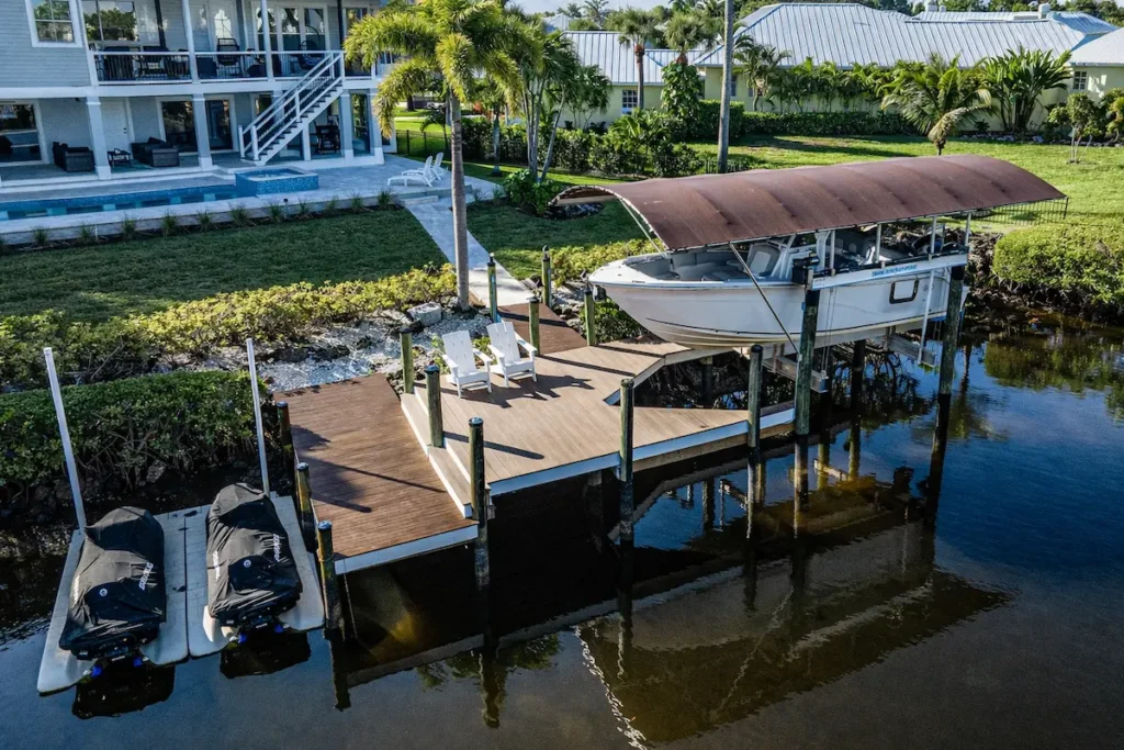 Aerial view of a waterfront home with a wooden dock, boat lift with canopy, and two jet skis beside a landscaped backyard with palm trees